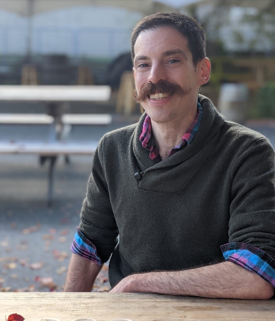 Man with a mustache sitting on a bench in a green sweater.